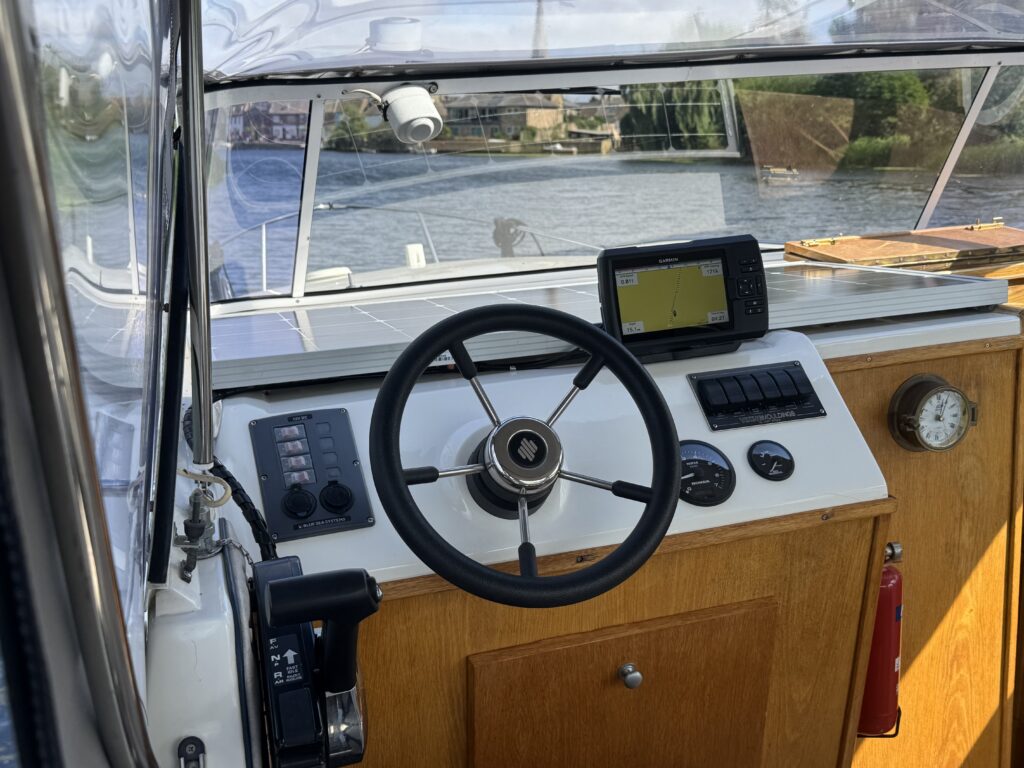 The helm of a boat featuring a wooden steering wheel, control panels with buttons, and a digital display. The view shows a waterway with trees and buildings in the background, along with visible sunlight reflecting on the water.