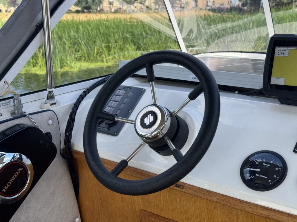 The interior of a boat featuring a black steering wheel with a silver emblem at the center. Nearby, there are control panels with switches and a digital display, alongside a round gauge for monitoring, all set against a light-colored dashboard.