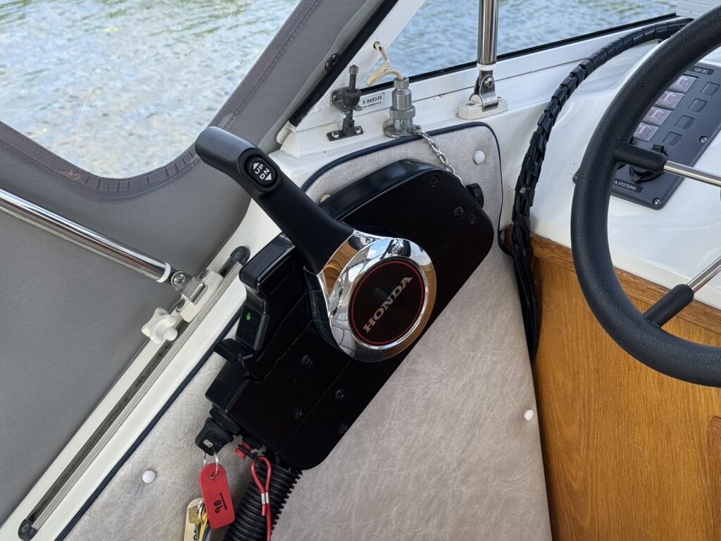 A black and chrome boat throttle control labeled 'Honda' is mounted on a panel near a window. The background shows a view of water and a black ropes coiled nearby on a wooden surface.