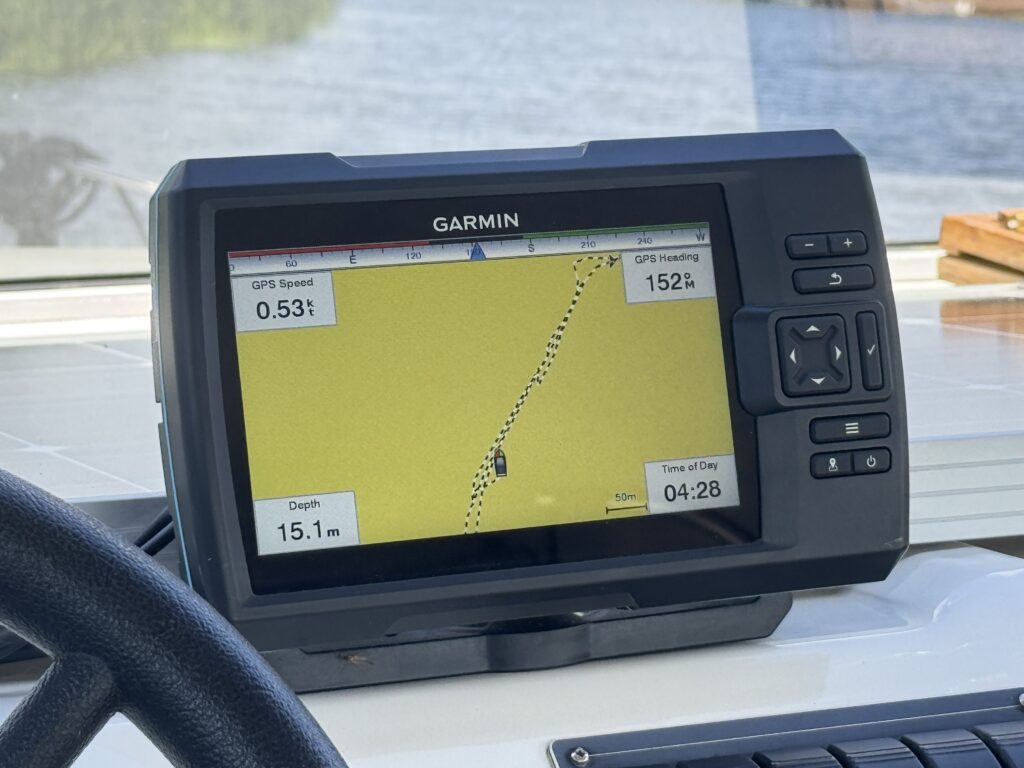 A Garmin GPS device displaying navigation information, including speed and depth readings. The screen shows a yellow map with a visible track line and a small boat icon, alongside data such as GPS speed, depth of 15.1 meters, and the time of day.