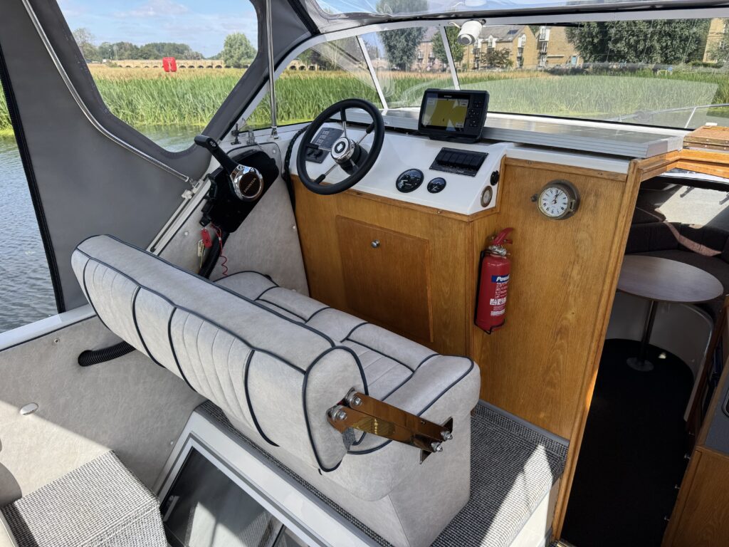 The interior of a boat features a white and gray striped captain's seat and a steering wheel in front of a dashboard with a display screen and gauges. A wooden panel includes a clock and a fire extinguisher, while the view outside shows a green landscape and water.