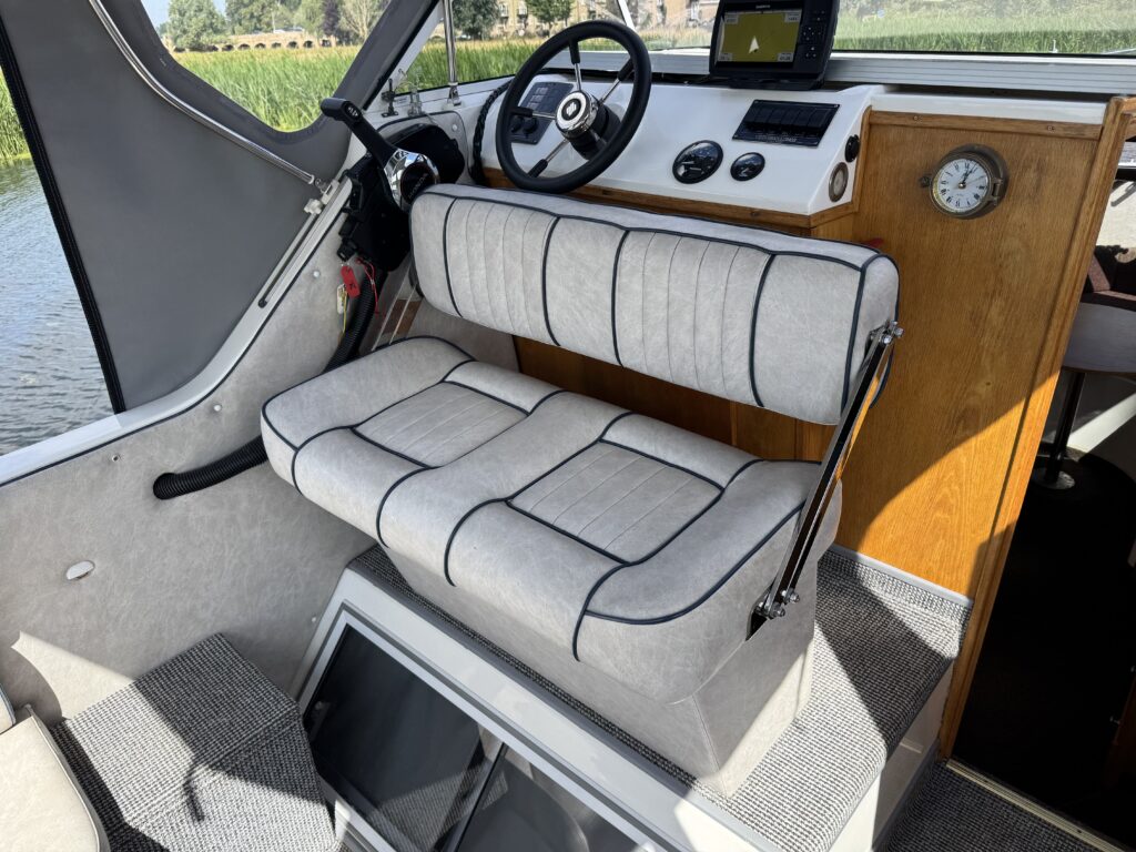 An interior view of a boat featuring a light gray upholstered bench seat with black piping. The seat is positioned near a steering wheel and dashboard with instrument gauges, while the background shows a window and a view of green vegetation outside.