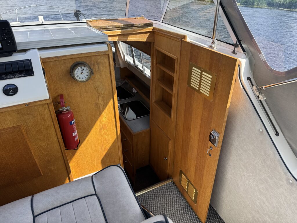 The interior of a boat features a wooden cabinet with a clock and a fire extinguisher. A small kitchen area is visible, including a sink. To the right, a door leads to a compartment, and sunlight shines through the windows, reflecting on the water outside.