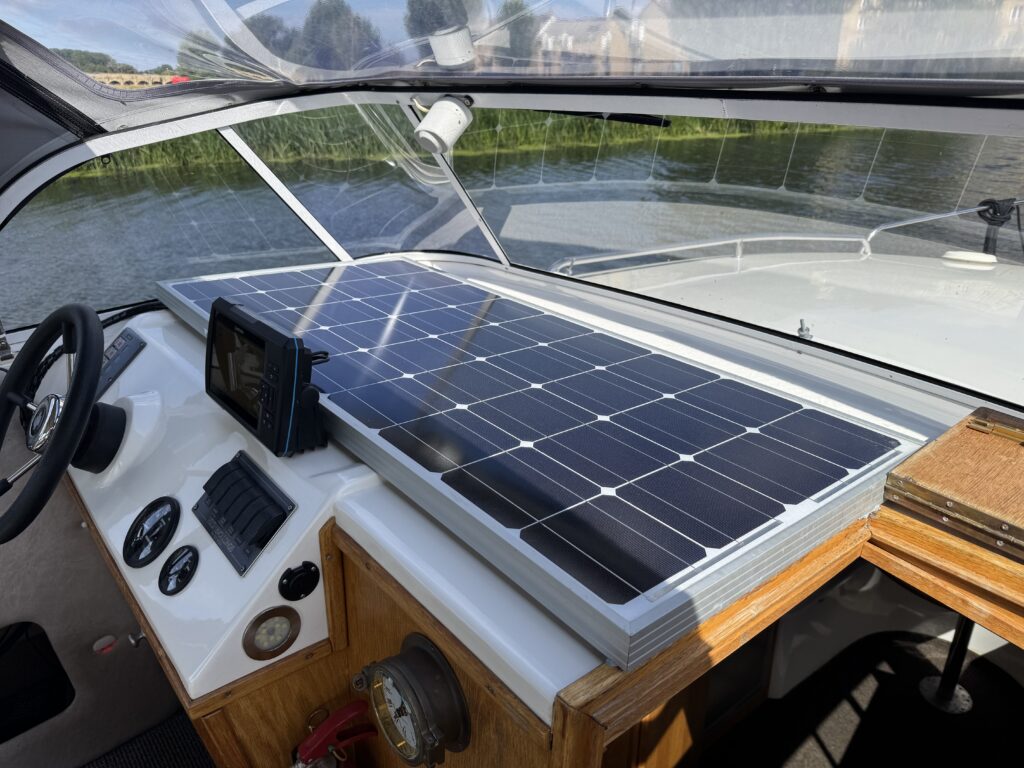 A close-up view of a boat’s cockpit featuring a solar panel mounted on the roof above the dashboard. The dashboard includes a steering wheel, control knobs, and a monitor, with natural light illuminating the space.