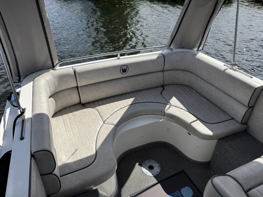 An L-shaped seating area in a boat, featuring light gray cushions and a textured surface. The background shows calm water and a partial canopy above, creating a shaded space.