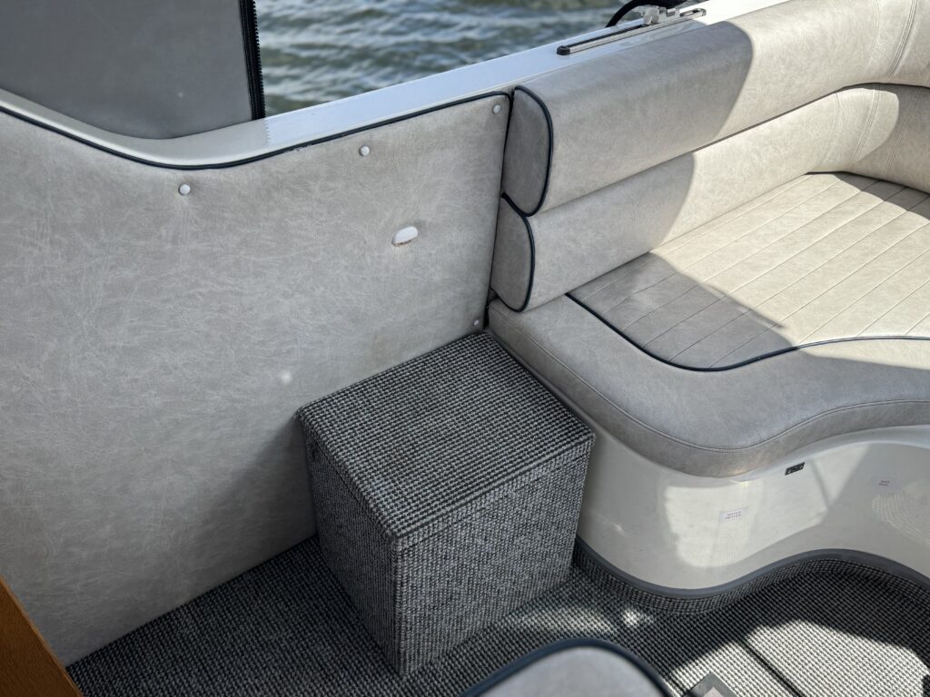 A corner of a boat featuring a light gray padded seating area and a square, textured storage box. The box sits on a patterned dark gray floor, with sunlight reflecting off the water through a nearby window.