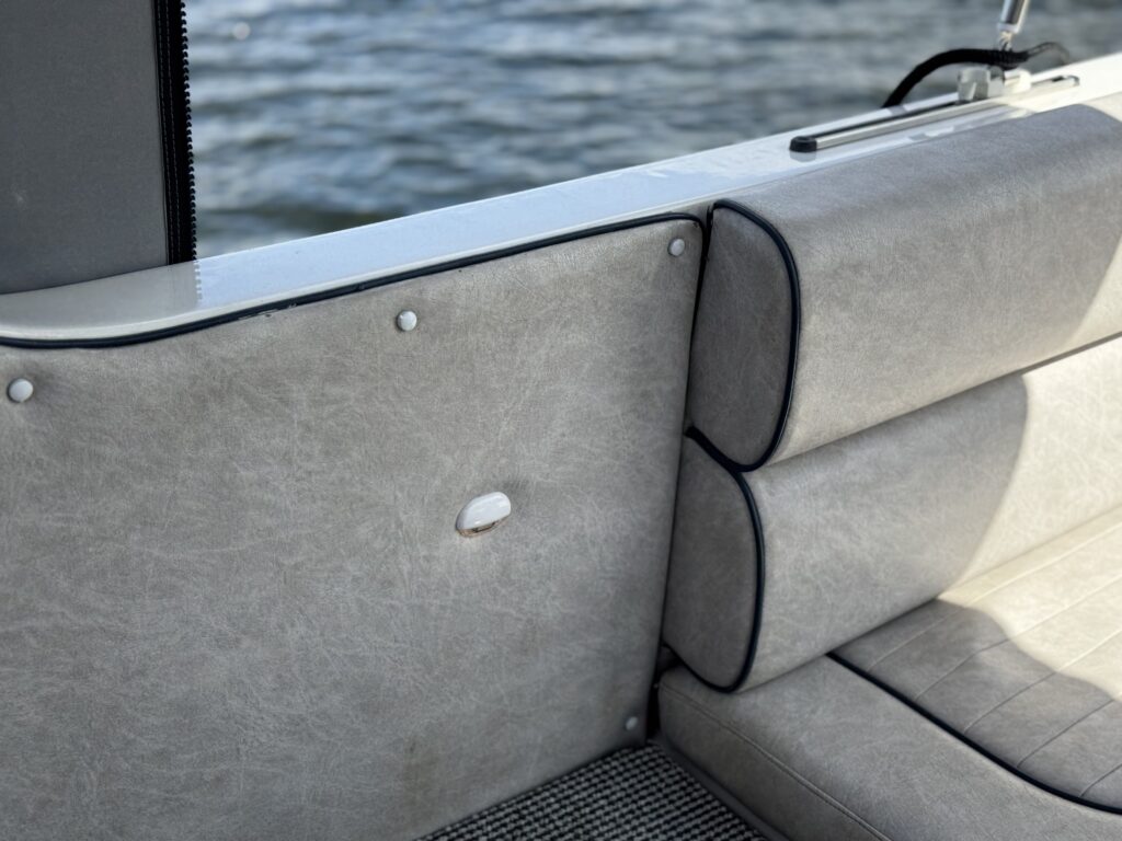 A close-up view of a light gray seating area on a boat, featuring a padded backrest and seat cushions. The textured fabric shows a seam along the edge, and the background displays calm water reflecting light.