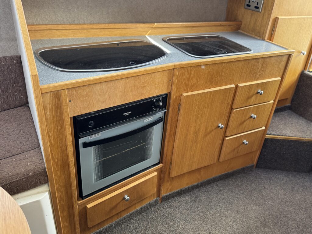 A compact kitchen area featuring a wooden cabinet with a built-in oven. The countertop has two black cooktops and a washing area, alongside several storage compartments including drawers and cabinets.