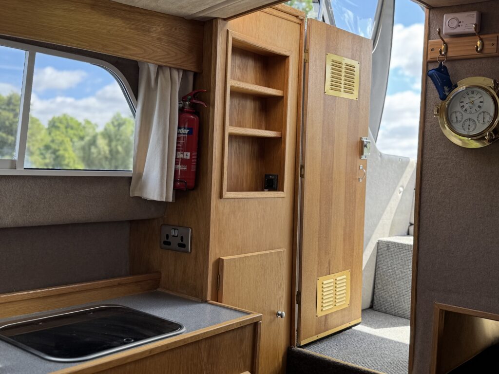The interior of a boat showing a kitchenette with a sink, wooden cabinetry, and an open door leading outside. A fire extinguisher is mounted on the wall, and there is a clock on the doorframe. Natural light streams through the window, illuminating the space.
