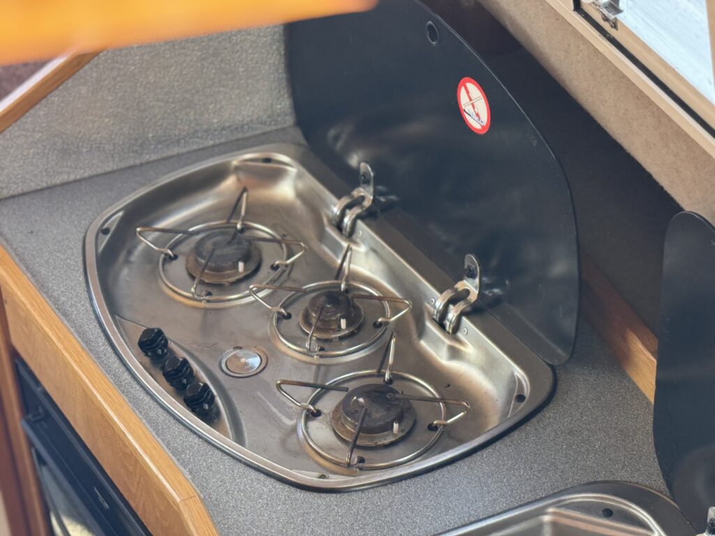 A compact stainless steel gas stove with three burners, mounted on a countertop. The stove has control knobs on the left side, and a fold-down cover is partially closed, revealing a textured gray surface underneath.