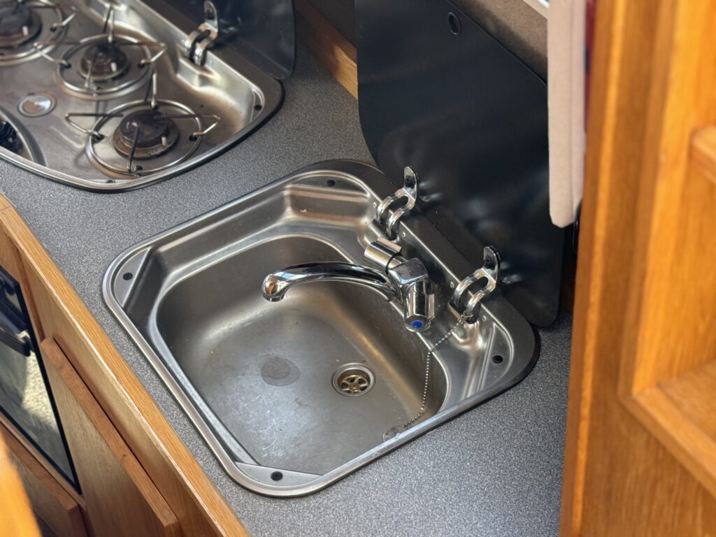A stainless steel sink with a single faucet is positioned in a countertop. To the left, there is a gas stove with two burners. The countertop has a smooth surface and is surrounded by wooden cabinetry.