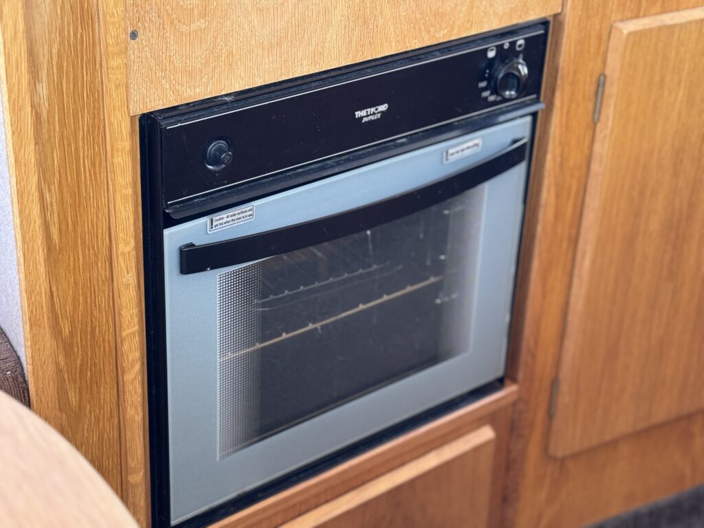 A black oven built into a wooden cabinet. The oven features a transparent door with a grid pattern and a knob for temperature control on one side. The surrounding area has warm wood tones, indicating a kitchen setting.