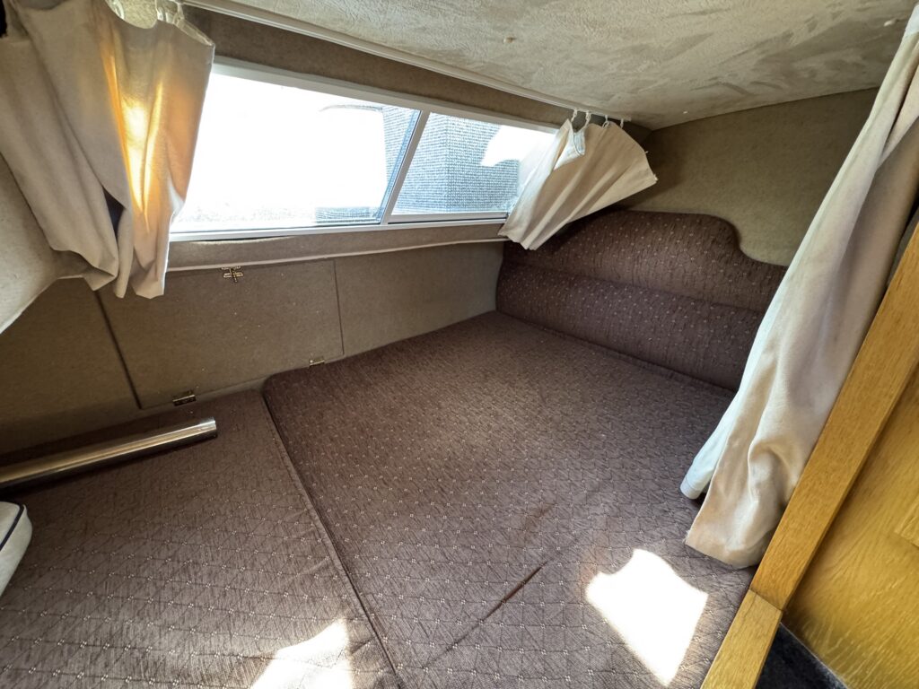 An interior view of a small sleeping area with a brown upholstered bed and a window with sheer curtains. Natural light filters through, illuminating the quilted bedspread and the sides of the wall, which are covered in beige fabric.