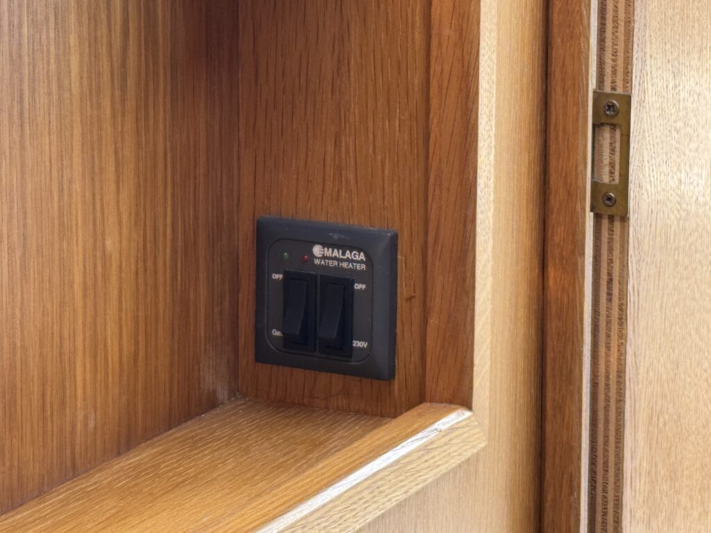 A black wall switch labeled "MALAGA WATER HEATER" mounted on a wooden panel. The switch has two positions labeled "ON" and "OFF" and is located in a wooden compartment, with a light wood grain visible around it.