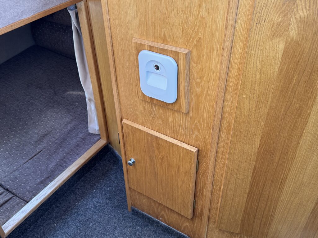 A close-up view of a wooden cabinet featuring a small gray electronic panel above a rectangular door. The door is secured with a metal knob, and there is a brown fabric cushion visible in the adjacent open compartment.