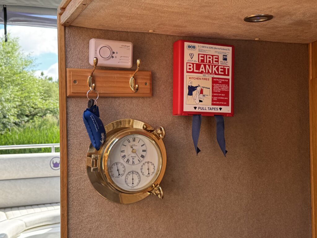 A round brass clock with multiple dials is mounted on a wooden panel alongside a red fire blanket box with a white label. A small device with buttons is attached above two hooks holding a keychain. The background features greenery visible through a window.