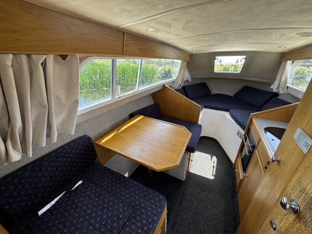 The interior of a small boat features a cozy seating area with blue upholstered cushions and a wooden table. Windows provide a view of greenery outside, while a compact kitchenette is visible on the right.