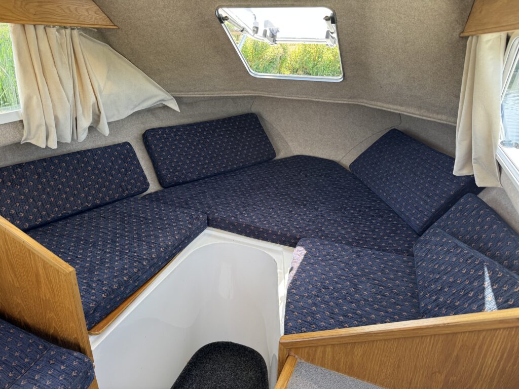 A cozy seating area in a small cabin with upholstered blue cushions arranged around a central table. Natural light enters through a triangular window, illuminating the space lined with light-colored curtains and wooden accents.