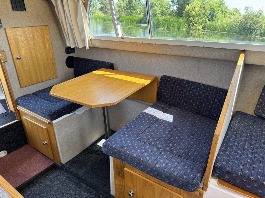 A cozy interior of a boat featuring a wooden table surrounded by upholstered seating. The table is positioned in front of two benches with blue fabric cushions, and a wooden cupboard is visible on the left side. Sunlight filters through the window, illuminating the space.