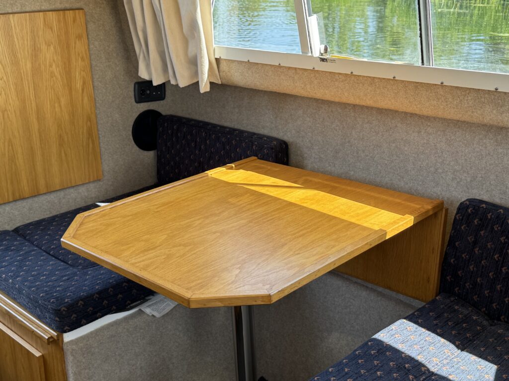 A wooden table with an angled edge is positioned between two upholstered benches in a cozy interior. The benches have a blue fabric with a subtle pattern, and a window is visible in the background, reflecting greenery and natural light.