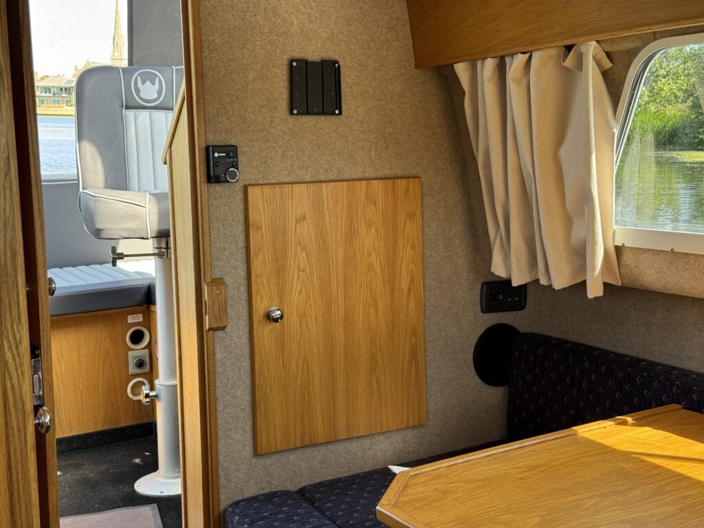 Interior view of a compact recreational space, showcasing wooden paneling, a table with dark upholstery, and a storage compartment on the wall. A window reflects natural light and offers a view of the water outside.