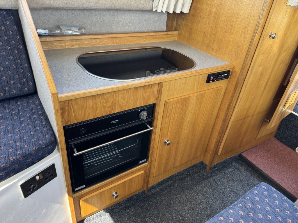 A wooden kitchenette featuring a built-in stove and a black oven beneath it. The countertop is gray, and the cabinetry has a natural wood finish. The background includes a patterned fabric seat and a doorway with a carpet.