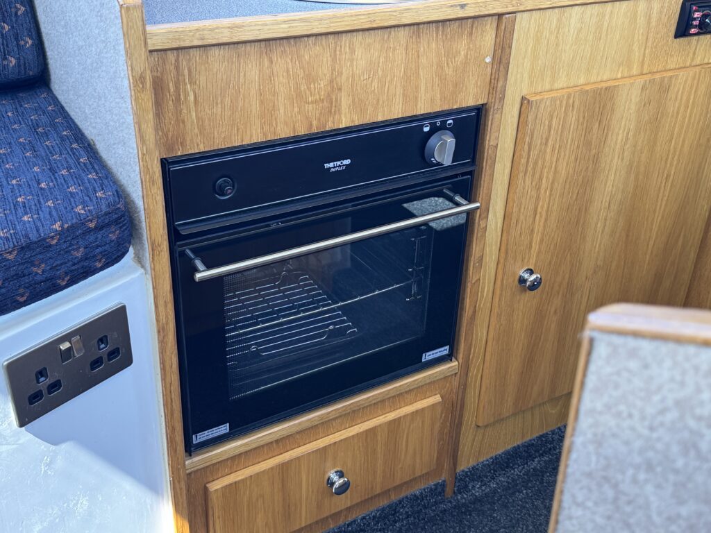 A black electric oven integrated into a wooden kitchen cabinet. The oven features a handle, control knob, and a glass door showing the inside rack. Below the oven, there is a small wooden drawer with a round handle.