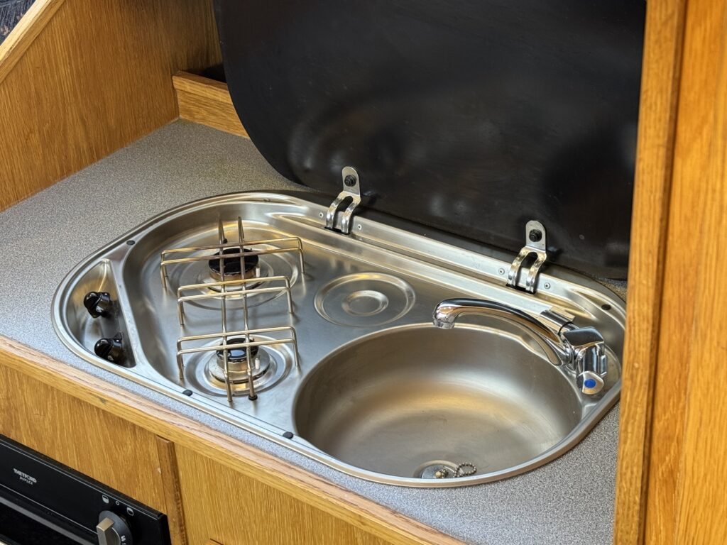 A stainless steel sink and a two-burner stove are mounted in a compact kitchen area. The sink features a faucet with a blue handle, and the stove has visible knobs and a protective cover placed above it.