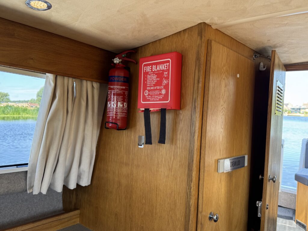 Inside a vessel, a wooden wall features a fire blanket mounted in a red case next to a fire extinguisher. A beige curtain hangs nearby, and a window shows a view of water and greenery outside.