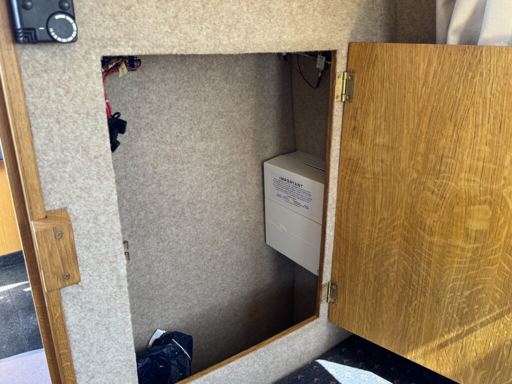 An open cabinet revealing a white electronic device along with wires connected to it. The cabinet's interior is lined with beige fabric, and the wooden door is partially closed. The floor shows a patterned surface and some shadowing from the cabinet.