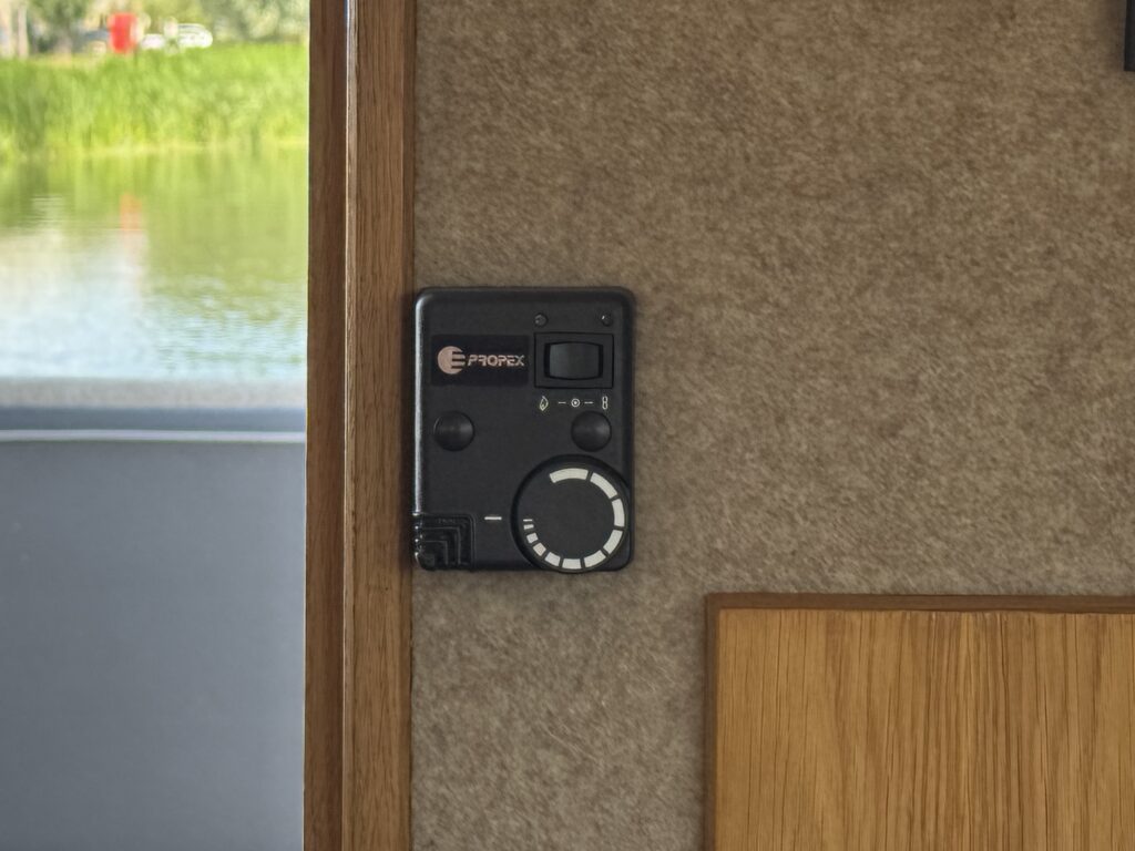 A black control panel mounted on a beige wall, featuring a circular dial, a power button, and an indicator light. In the background, there is a body of water with lush greenery visible outside.