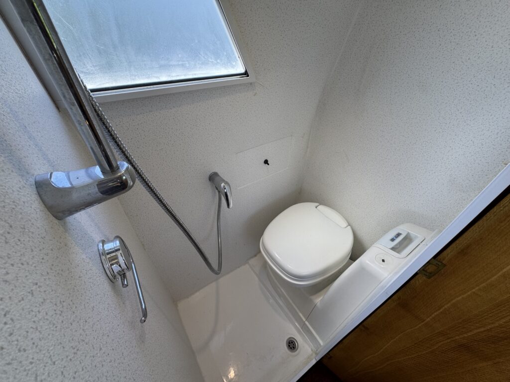 A small bathroom space featuring a modern white toilet next to a shower with a handheld showerhead. The wall is textured white, and there is a small window providing natural light.