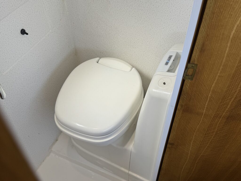 A white toilet with a closed lid is positioned in a small bathroom corner. The walls are light-colored, and there is a wooden door partially visible on the right side of the image.