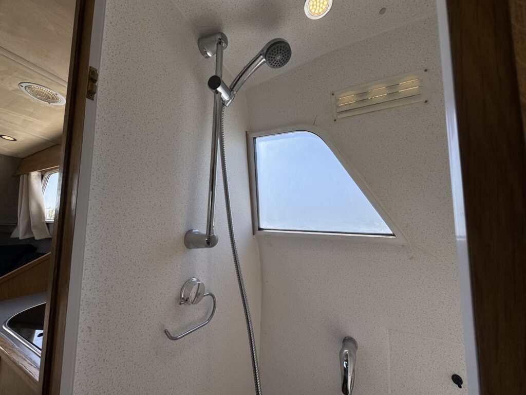 A compact shower area with a handheld showerhead mounted on the wall. A small window is visible, providing natural light, and the interior walls are finished in white with a speckled texture. The floor includes a drain.