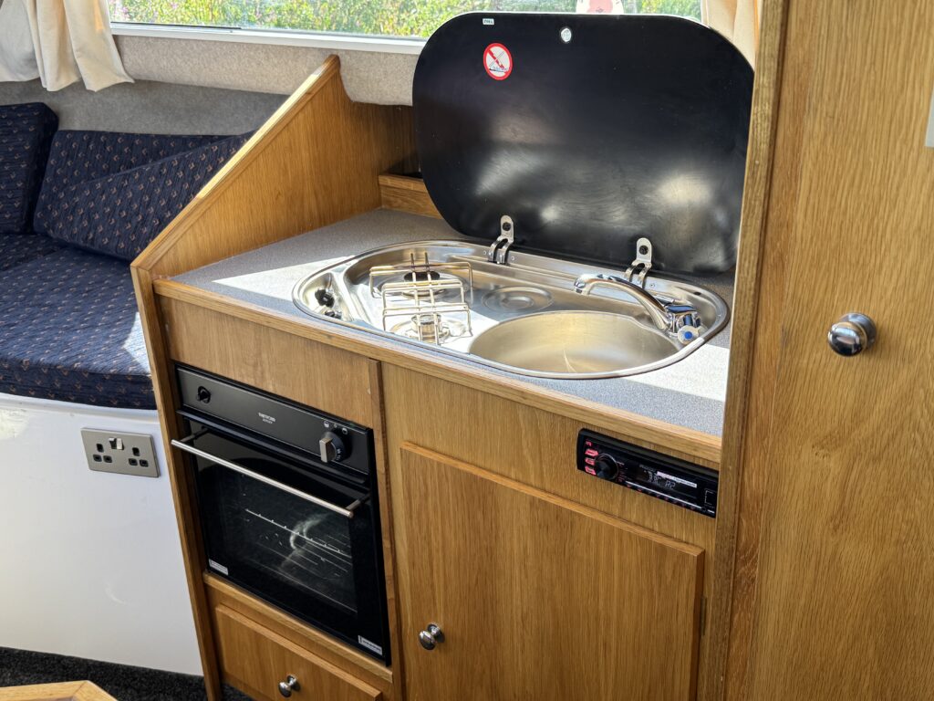 An interior view of a kitchen area in a caravan, featuring a compact stove and sink with a black lid. Below, there is an oven with a black front panel. The cabinetry is brown wood, and there is a seating area visible in the background.