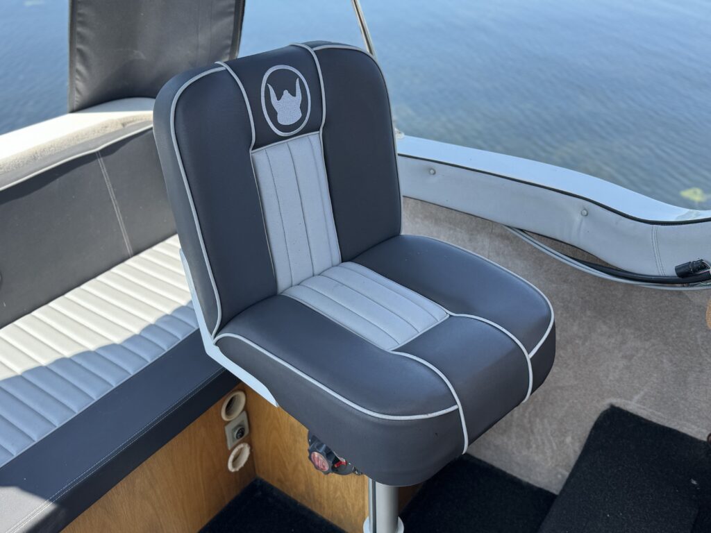 A gray and white boat seat with a high backrest and a logo at the top, positioned on a bright wooden surface. The background shows calm water and a bright sky.
