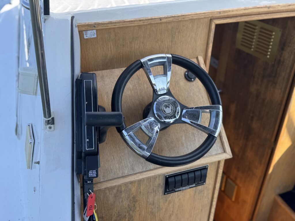 A close-up view of a boat's steering wheel, mounted on a wooden console. The steering wheel is chrome and black, and there is a control panel nearby with switches and a key attached by a yellow lanyard.