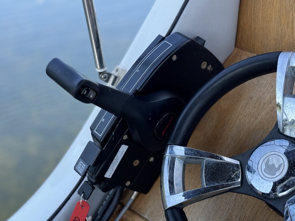 A close-up view of a boat's control panel featuring a throttle lever on the left, with a key attached, and a steering wheel on the right. The background displays calm water reflecting light.