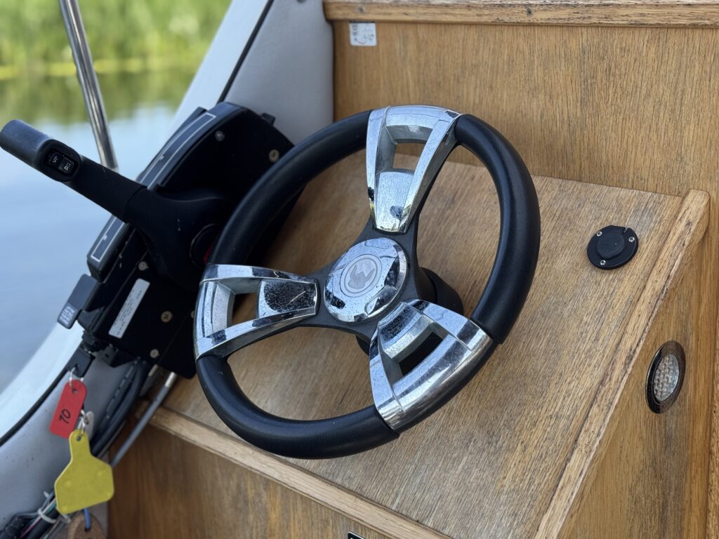 A chrome steering wheel with a black grip is positioned on a wooden boat dashboard. To the left, there is a throttle control and various buttons, with colored tags hanging nearby and a view of foliage reflected in the water outside.