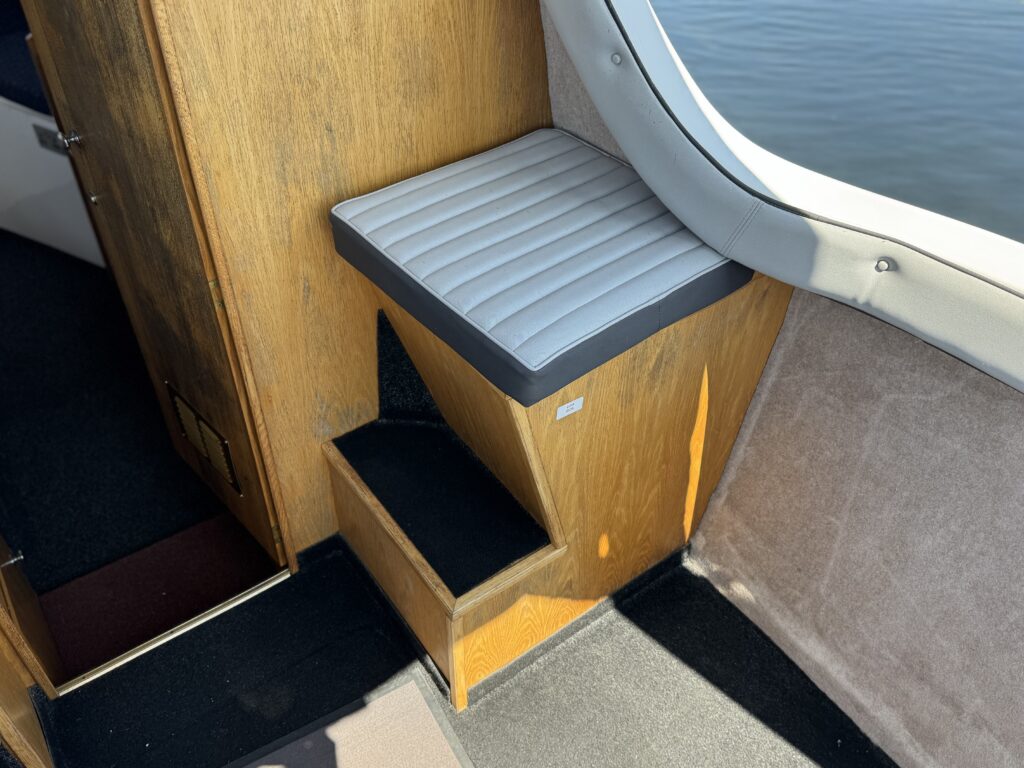 An interior corner of a boat featuring a wooden wall with a small cushioned seating area and a step down to the floor. The seat has a gray cushion with a ribbed pattern, while the background shows water through a large window.