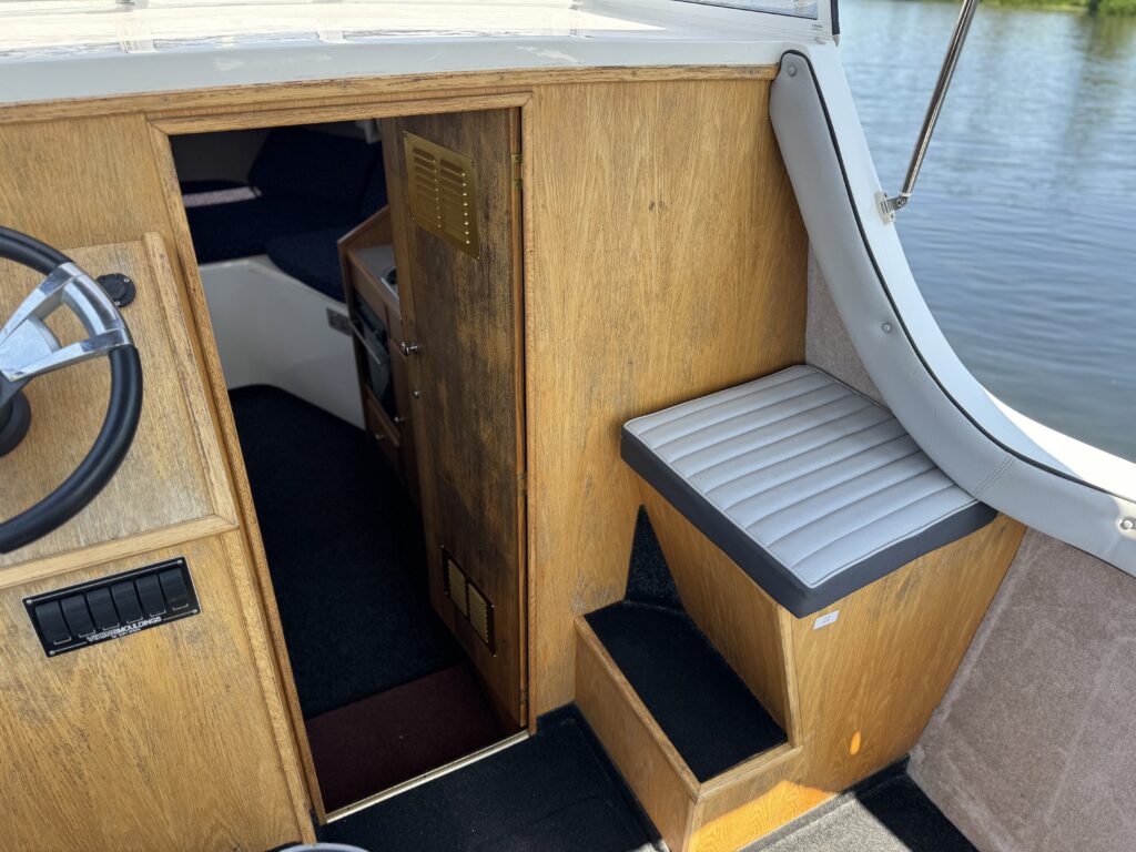 The interior of a boat features a steering wheel on the left and a wooden door entrance on the right leading to another cabin area. A padded bench seat is located beside the door, while the floor has dark carpeting and natural light reflects off the water outside.