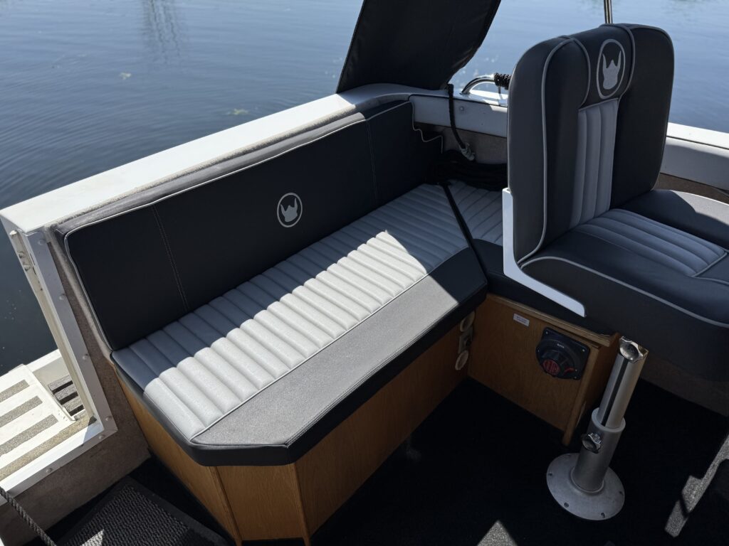 A corner seating area on a boat, featuring gray and white padded upholstery with a wooden base. The scene includes a captain's chair with a black and gray design and reflections on the water in the background.