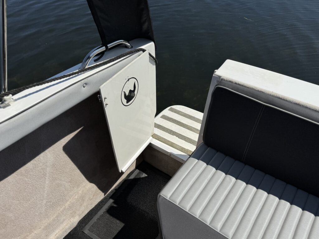 A close-up view of a boat's interior showing a white door with a black silhouette of a fish. The boat's seating is gray with vertical lines, and part of a striped boarding platform is visible, along with dark water in the background.