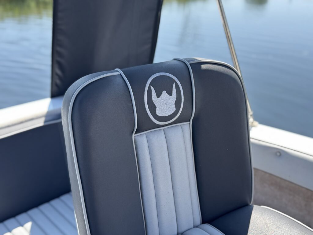 A close-up view of a boating seat with black and gray upholstery, featuring a logo with a crown design on the headrest. The background shows calm water reflecting the sunlight.