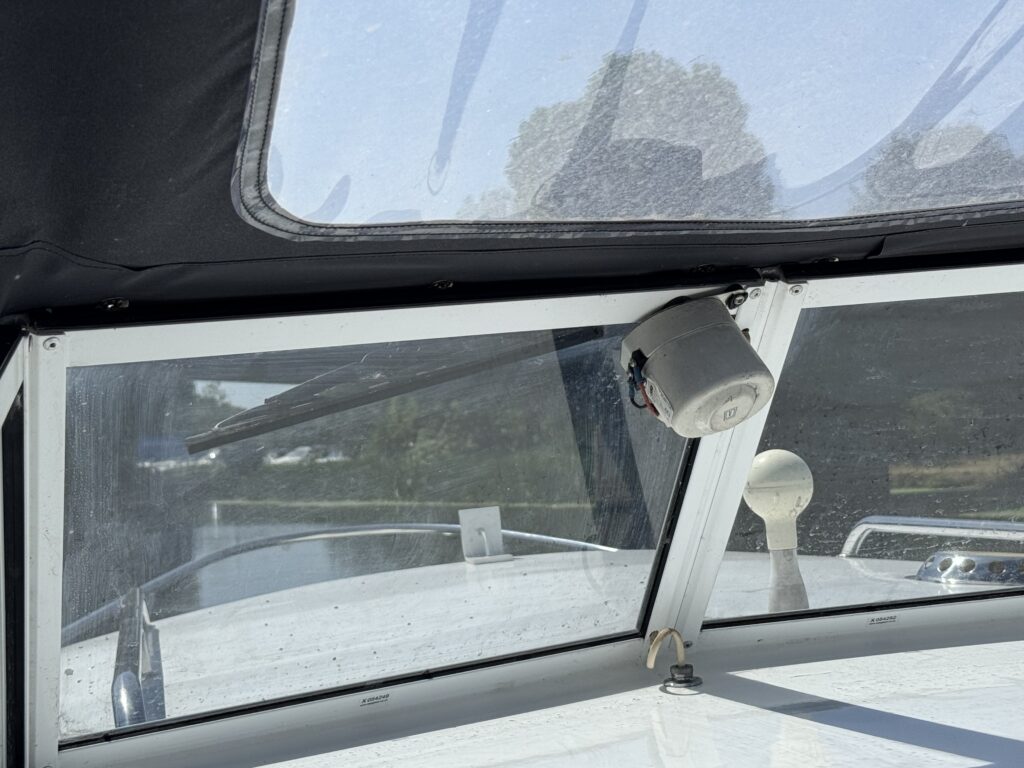 A view from inside a boat through a large windshield, showing the dashboard with a steering wheel and a compass. The scenery outside includes green trees and water, indicating a calm day on the water.