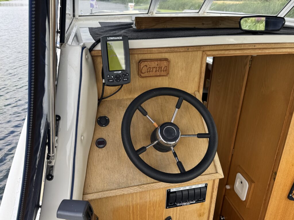 A marine steering wheel mounted on a wooden dashboard, with a digital navigation display attached above it. The dashboard is made of light-colored wood and includes several buttons and controls, with a view of water visible outside the window.
