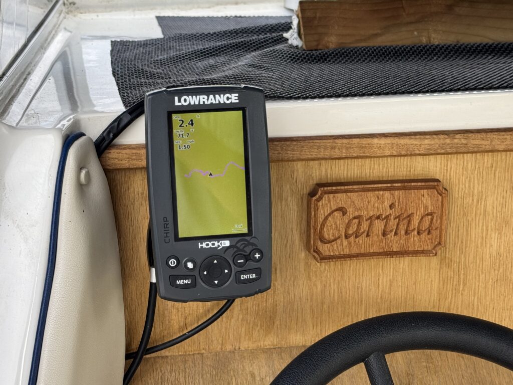 A Lowrance fish finder displayed on the dashboard of a boat, showing a map with navigation details. Next to it is a wooden plaque engraved with the name 'Carina' on the side of the boat.