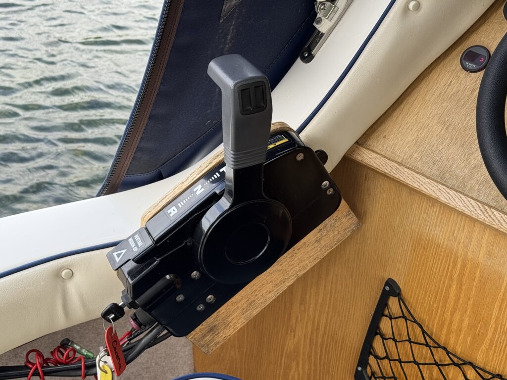 A close-up view of a boat throttle control mounted on a wooden surface. The control has a black lever in the upright position and includes numerical indicators for throttle settings. In the background, water is visible through a window.