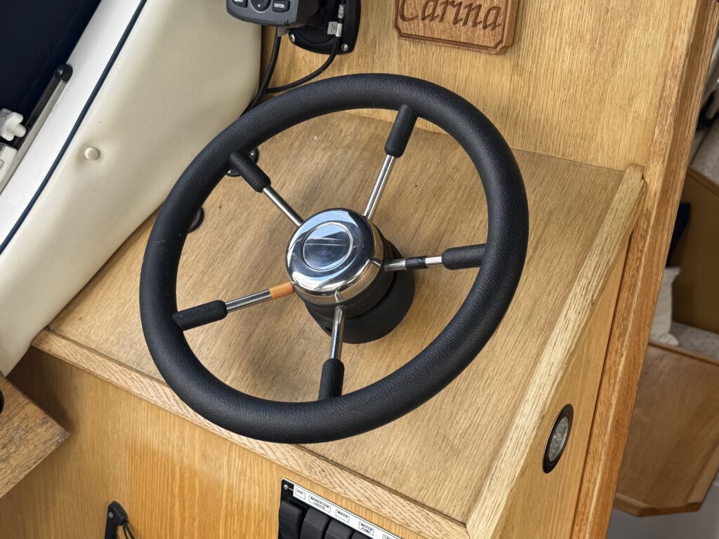 A steering wheel with a black rubber grip is mounted on a wooden dashboard. The dashboard features a beige wood finish and includes a small control panel with switches beneath the wheel. A nameplate reading "Carina" is visible on the side.