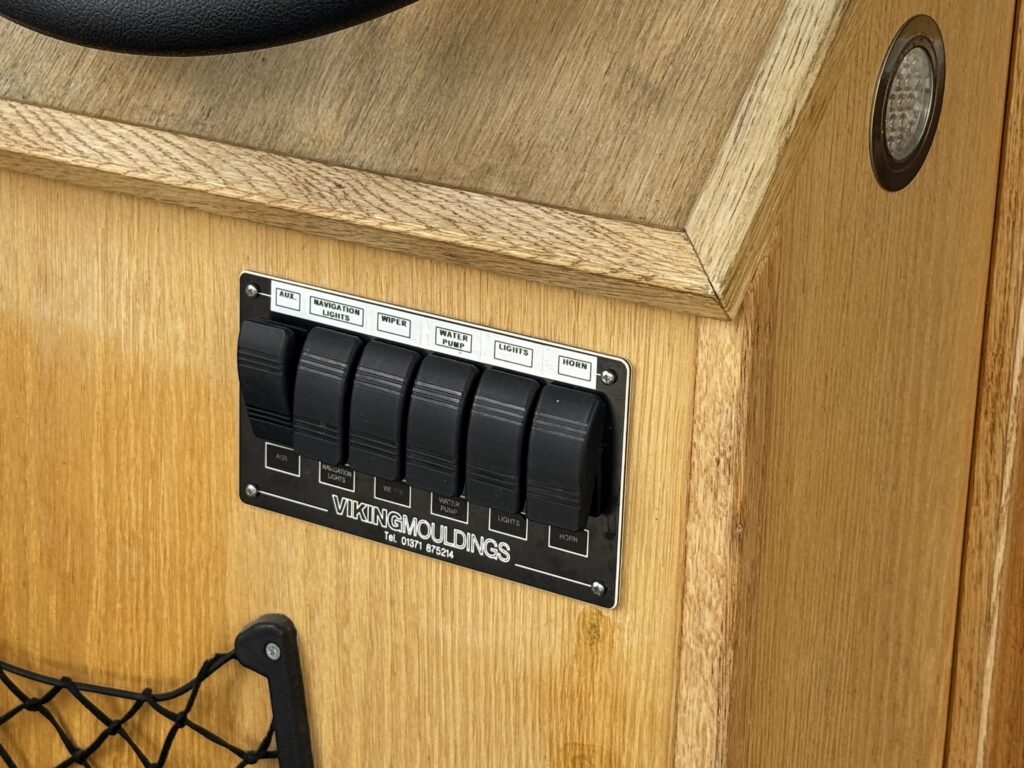 A close-up view of a wooden surface featuring a control panel with six black toggle switches labeled "Wake," "Power," "Lights," "Transom," "Cabin," and "Down". The panel is mounted on an oak background with a textured finish.